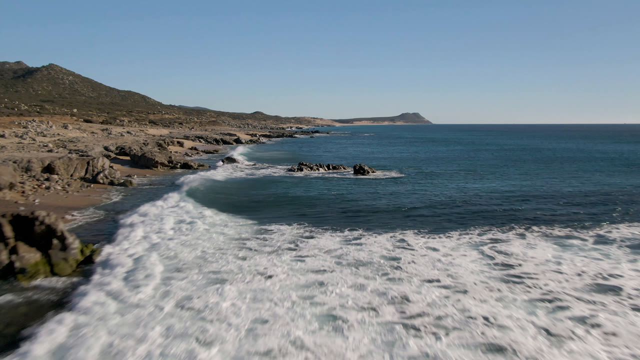 Opening frame of Laguna Beach Los Cabos, Baja California Sur, with white surf rolling over foreground water, dark rocks near shore, dry hills at left, and no visible people.