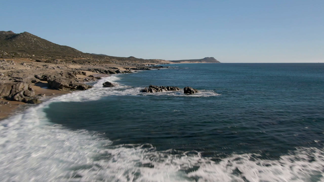 Laguna Beach Los Cabos coastline with a sandy cove on the left, dark rock outcrops in the water, a low hill behind the shore, and no visible people.