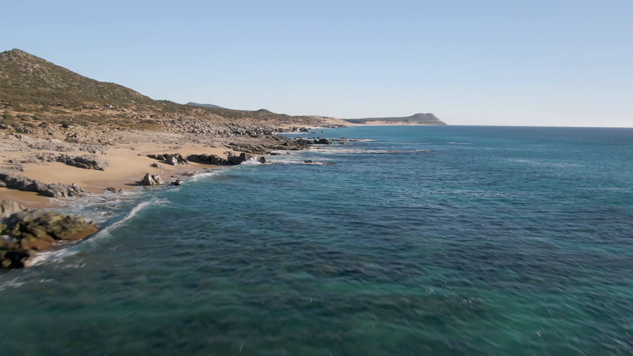 Opening aerial view of Laguna Beach Los Cabos with blue-green ocean in the foreground, tan sand and dark rocks at left, low hills behind, and no visible people.