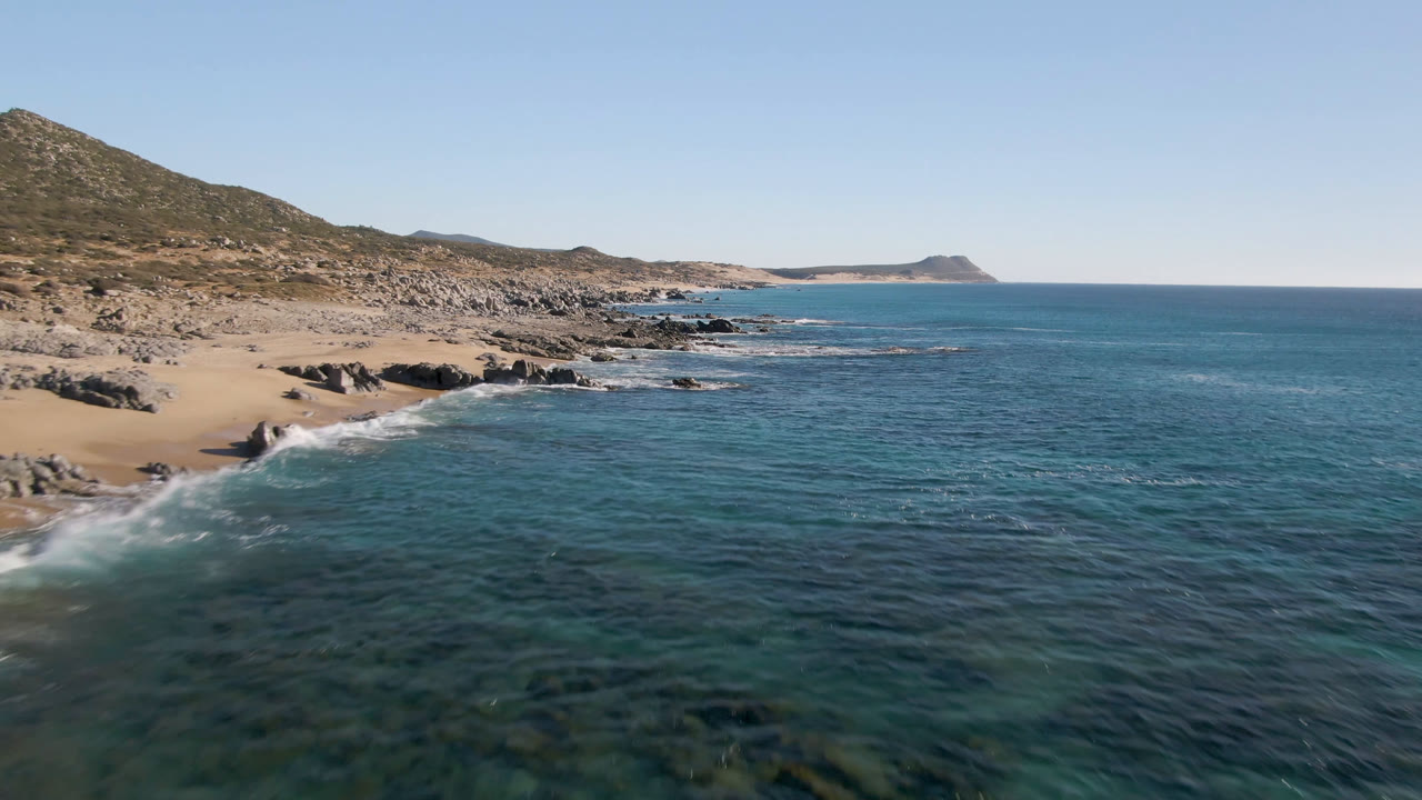 Aerial frame over Laguna Beach Los Cabos showing white surf curling onto rocks and sand at left, clear blue water across the foreground, distant coastline, and no visible people.