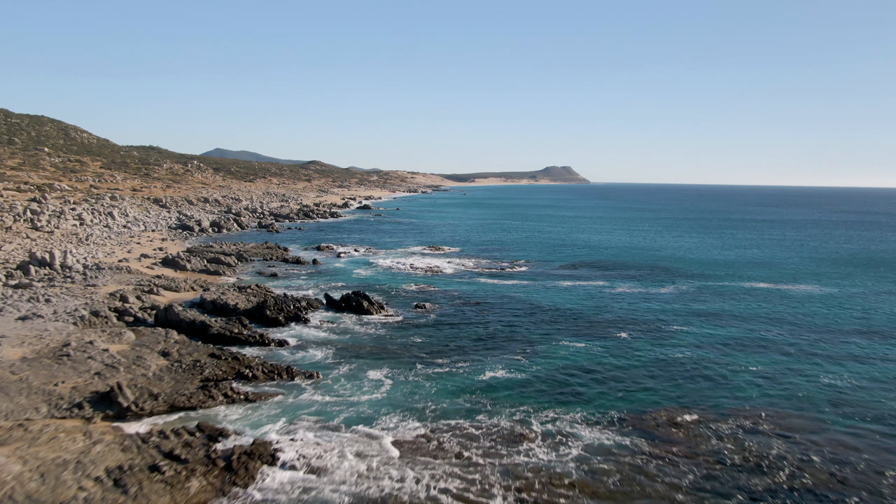 Aerial view of Laguna Beach Los Cabos with jagged rocks along the left shoreline, blue-green water to the right, white surf in the foreground, and no visible people.