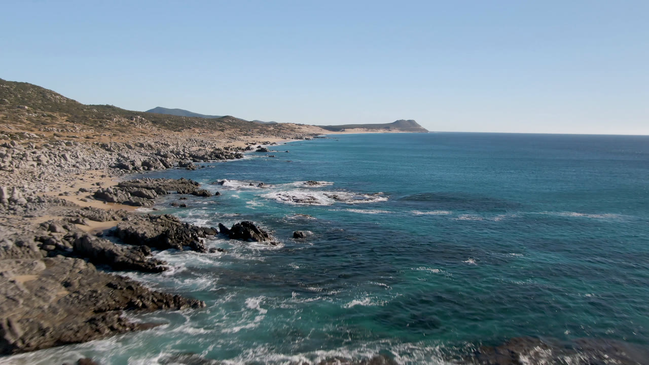 Laguna Beach Los Cabos shoreline seen from above, with dark rocks breaking the turquoise shallows, tan hills along the left, a distant headland on the horizon, and no visible people.