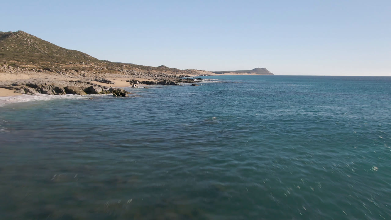 Laguna Beach Los Cabos shoreline continues along the left side of the frame, with calm blue water across the foreground, a distant headland, and no visible people.