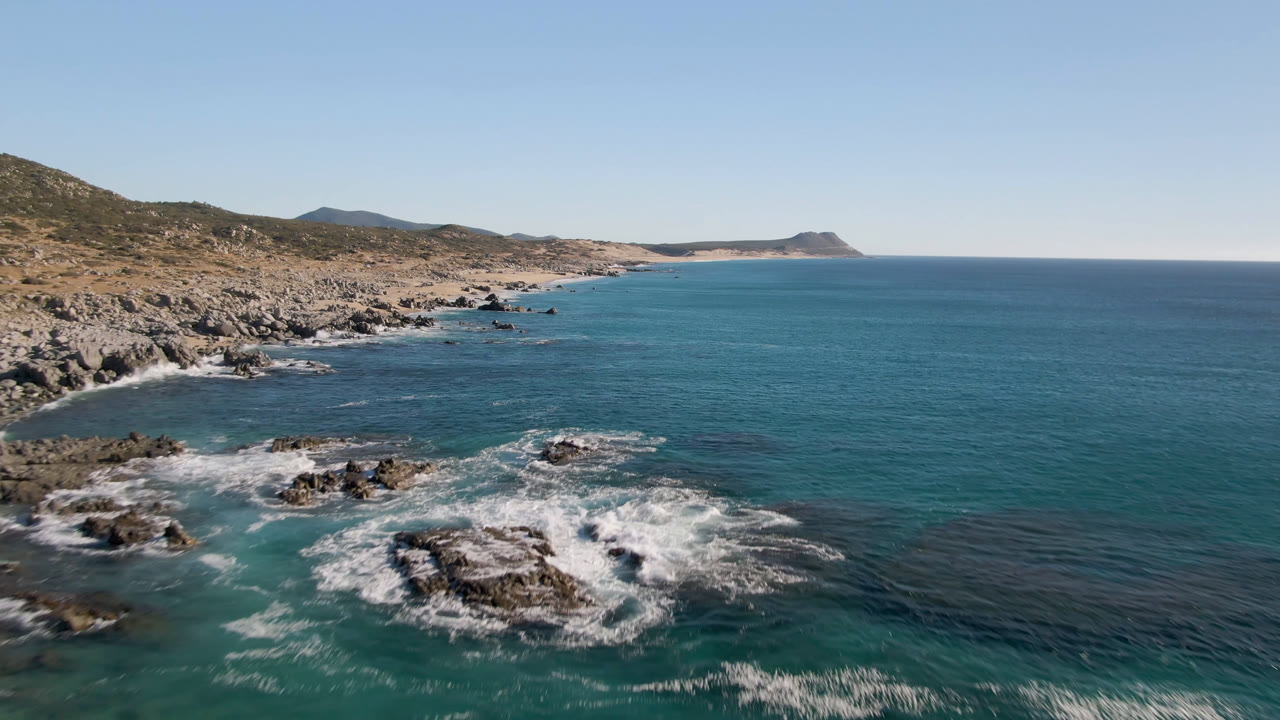 Aerial view over Laguna Beach Los Cabos, Baja California Sur, Mexico, showing white surf around foreground rocks, dry hills on the left, open blue water on the right, and no visible people.