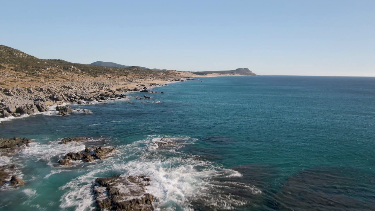 Aerial frame from Laguna Beach Los Cabos, Baja California Sur, Mexico, with foamy waves spreading across dark rocks, the arid shoreline receding left to right, and no visible people.