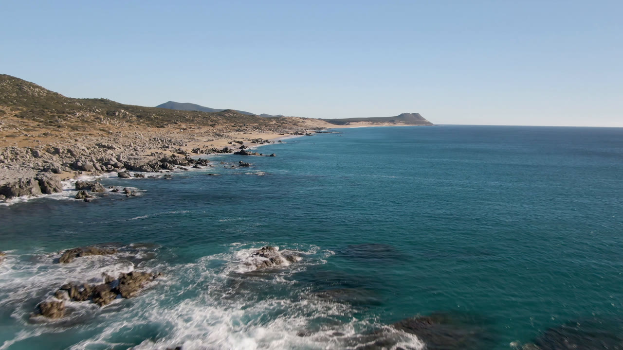 Wide coastal aerial with rocks, blue water, and a distant headland.