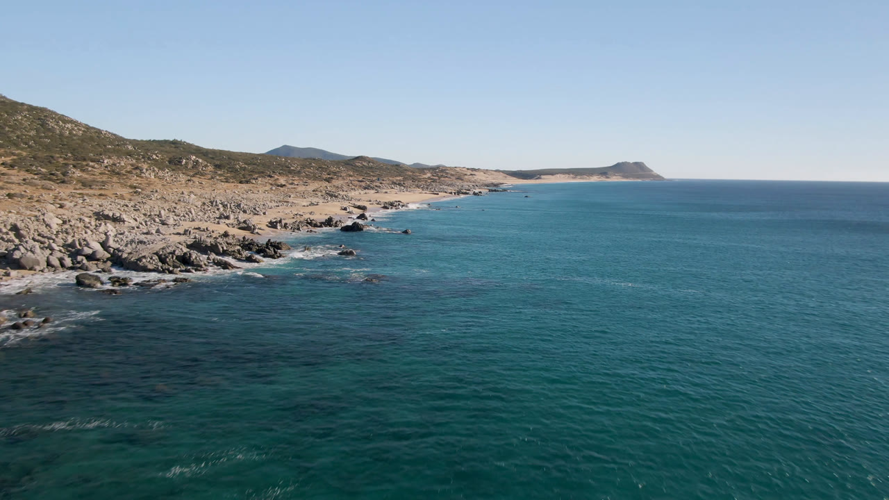 Opening aerial frame of Laguna Beach Los Cabos with jagged rocks on the left, turquoise ocean on the right, distant hills on the horizon, and no visible people.