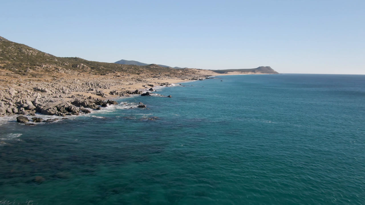 Aerial view one second in at Laguna Beach Los Cabos, with foamy water along the rocks, a broad open ocean area, scrubby hills, and no visible people.