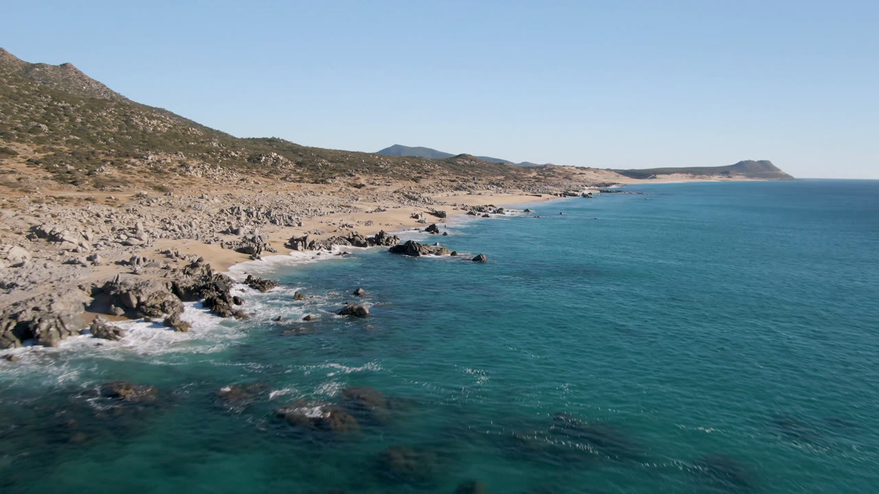 Opening frame of Laguna Beach Los Cabos with turquoise water on the right, dark shoreline rocks below, tan beach and scrub hills beyond, and no visible people.