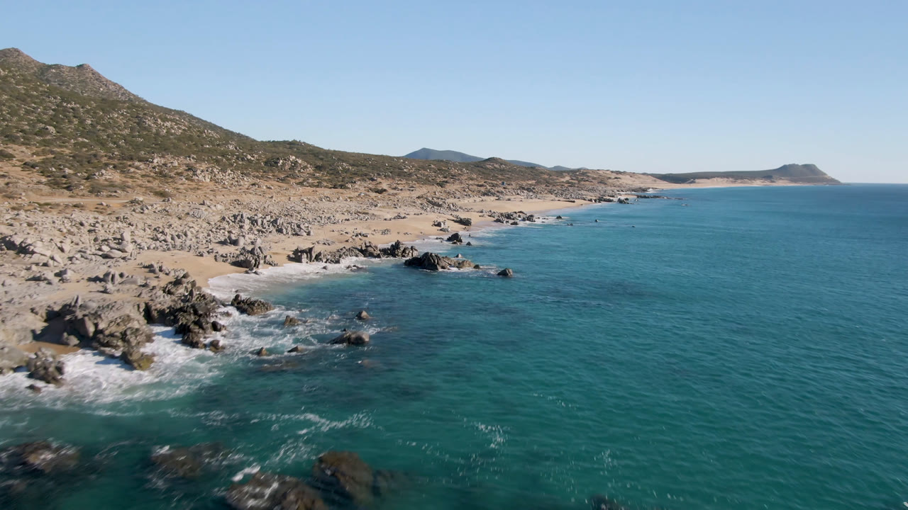 Second frame of Laguna Beach Los Cabos showing white water curling around nearshore rocks, a narrow sand strip, dry hills, distant coast, and no visible people.