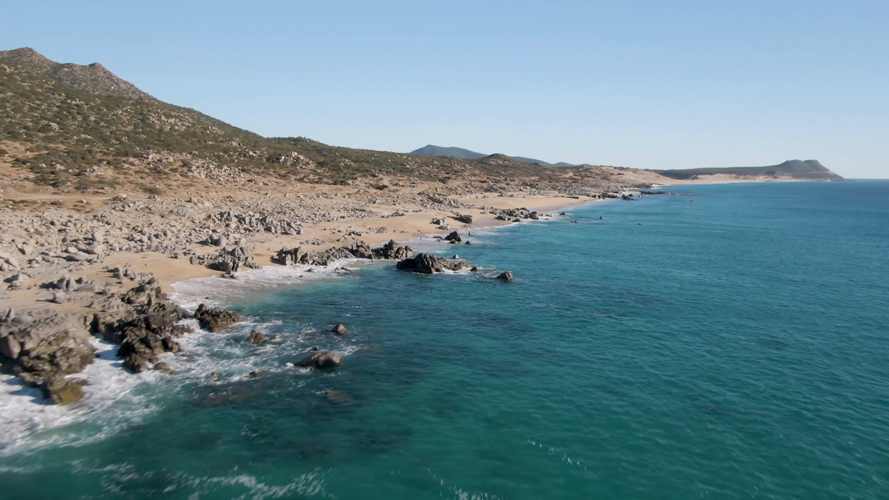 Wide aerial of rocky beach, clear ocean, and low coastal hills.