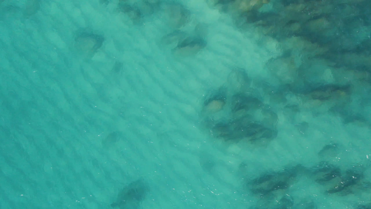 Overhead view of turquoise water at Laguna Beach Los Cabos, Mexico, with rippled sand patterns, scattered dark submerged rocks, and no visible people.