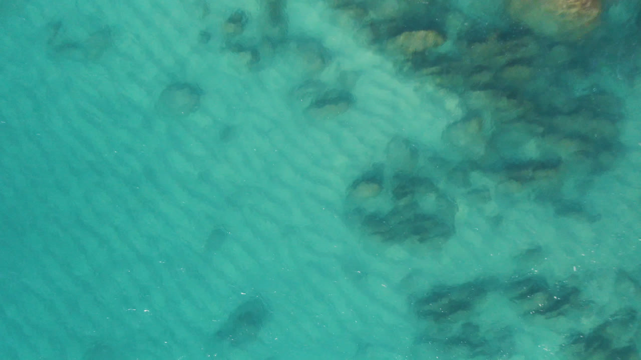 Aerial frame from Laguna Beach Los Cabos, Mexico, showing aqua shallows, fine sand ripples, dark underwater rock patches, and no visible people.