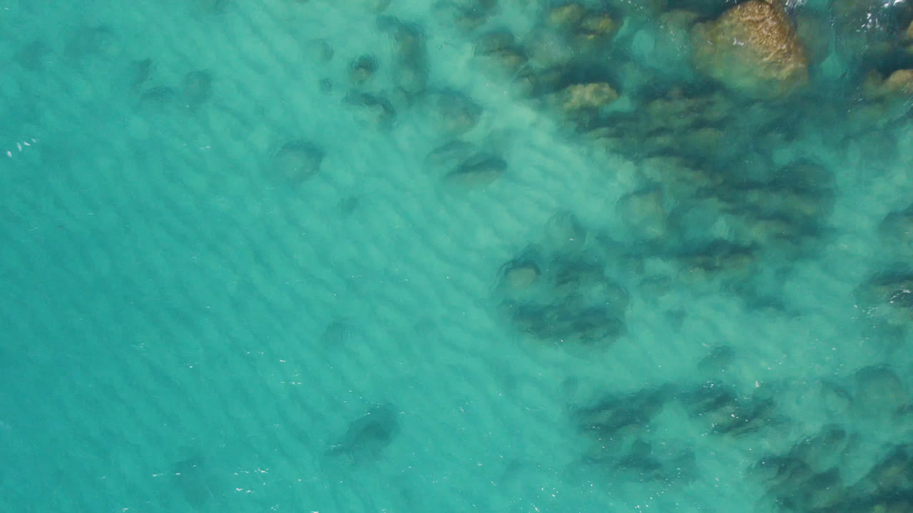 Turquoise water with submerged rocks and pale coastal boulders near the upper edge.