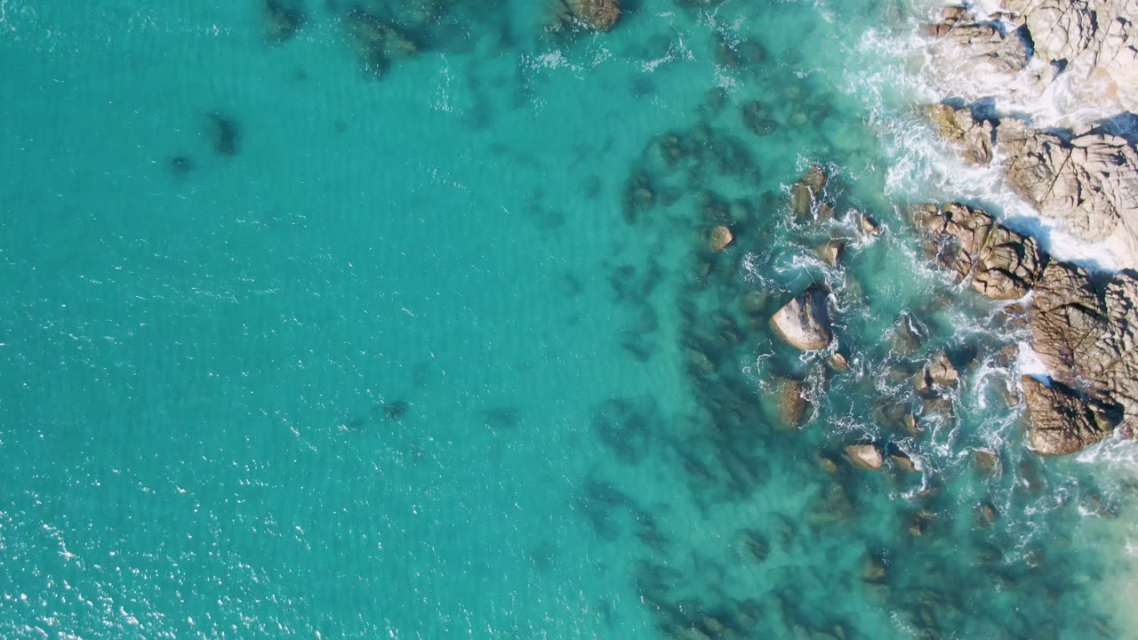 Top-down view of turquoise water and rocky shoreline at Laguna Beach Los Cabos, with white surf around the rocks and no visible people.