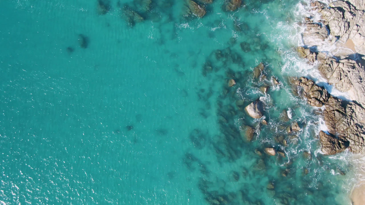 Aerial frame over Laguna Beach Los Cabos showing open turquoise water, pale shoreline rocks, darker underwater patches, and no visible people.