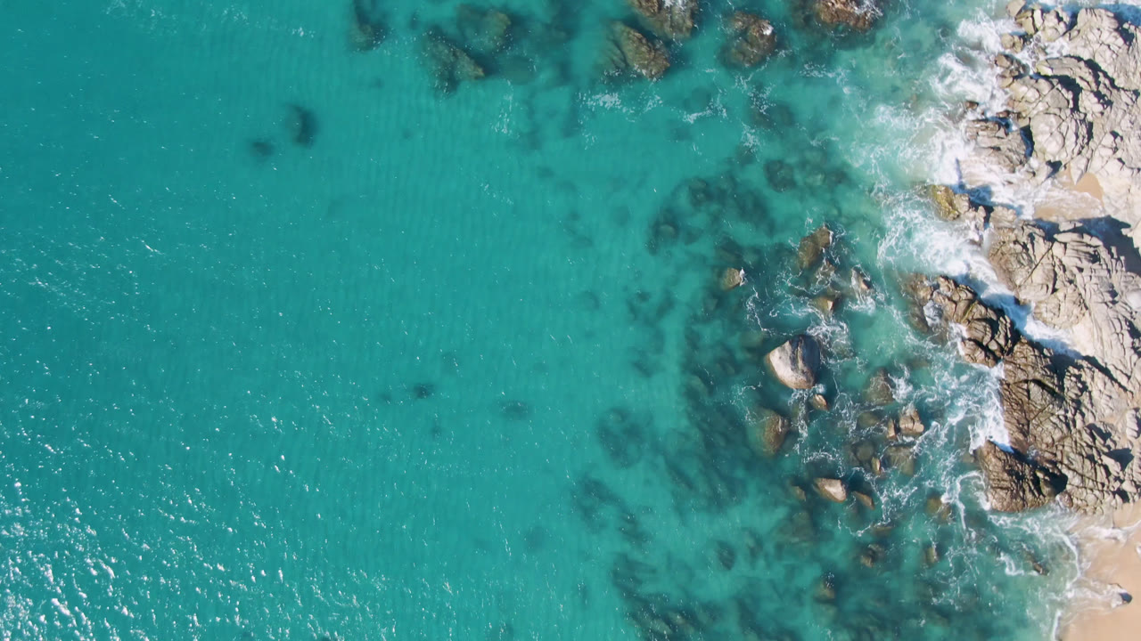 Top-down Laguna Beach Los Cabos view with turquoise shallows, clustered rocks near the coast, white foam, and a narrow sandy edge with no visible people.