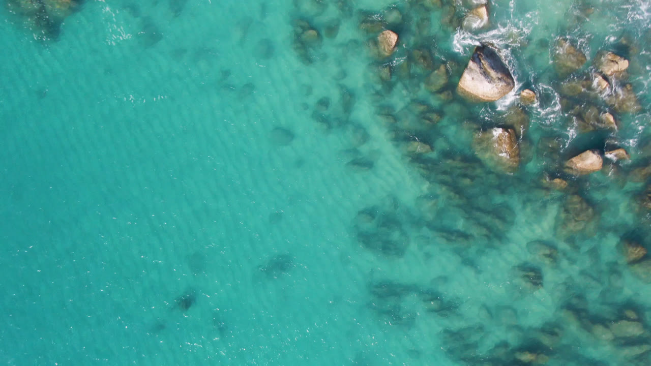 Overhead view of turquoise water at Laguna Beach Los Cabos with tan rocks near the top right, faint submerged rock shapes, white surf, and no visible people.