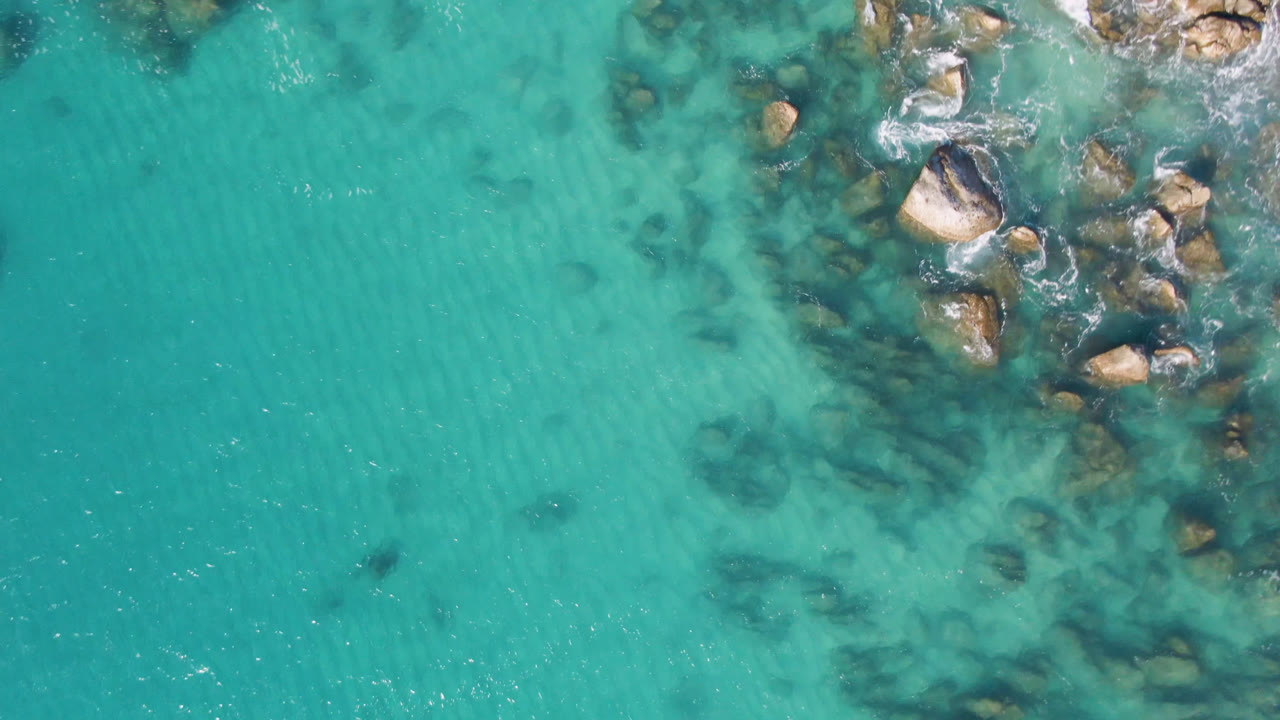 Aerial frame from Laguna Beach Los Cabos showing broad turquoise water, a dense cluster of shoreline boulders in the upper right, scattered submerged rocks, and no visible people.