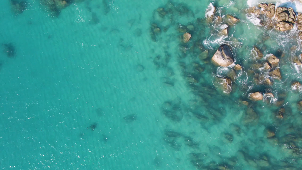 Turquoise ocean surface with rocks and foamy wave wash on the right.