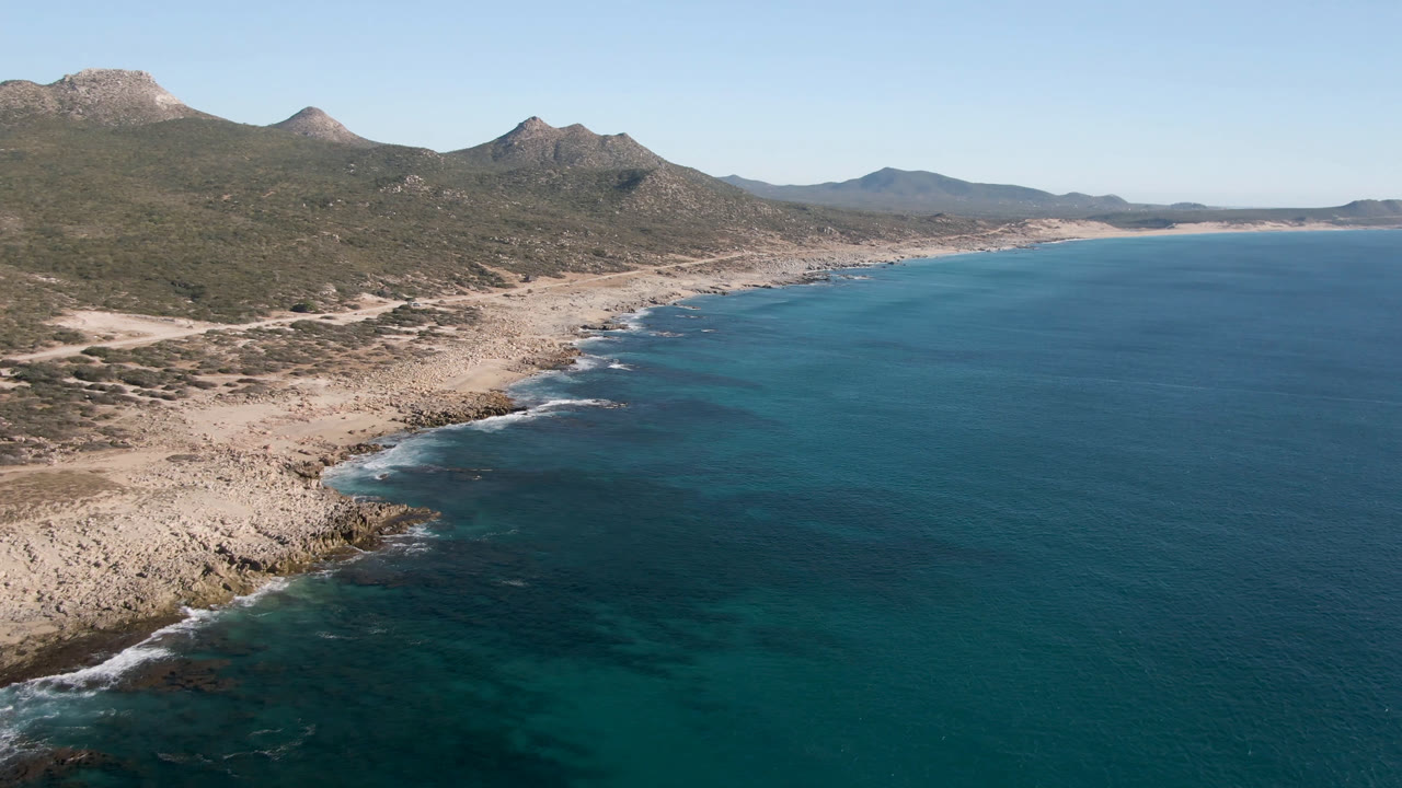 Aerial frame of Laguna Beach Los Cabos showing turquoise water along a rocky shore, scrub-covered land, distant mountains, and no visible people.