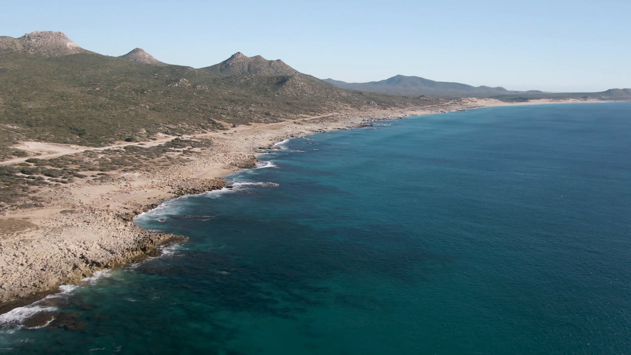 Wide aerial coast with open ocean, rocky shoreline, scrubland, and mountain ridge.