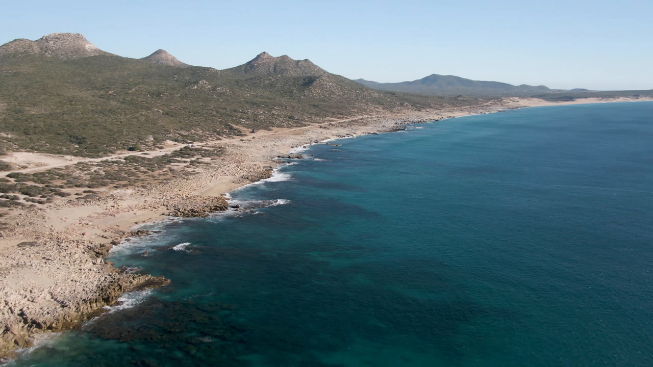 High aerial frame over Laguna Beach in Los Cabos, Mexico, with turquoise water along a pale rocky shore, scrubland, mountain ridges, and no visible people.