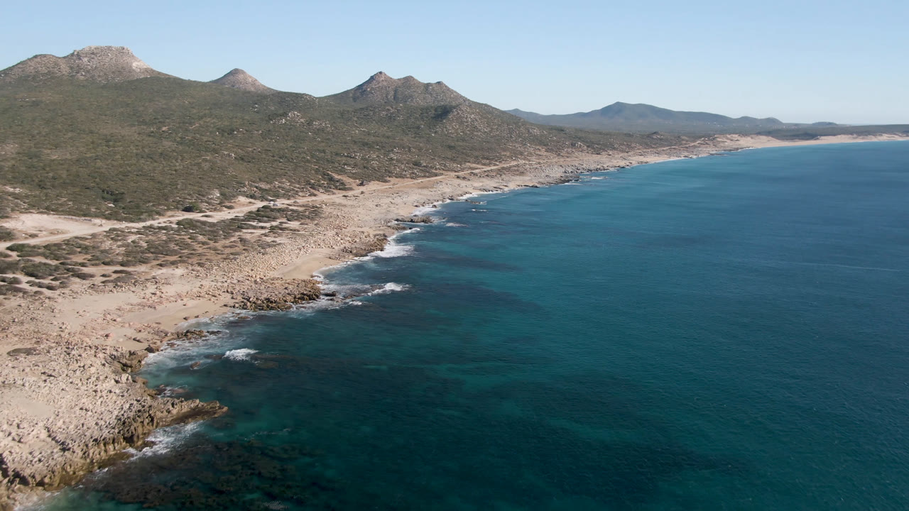Aerial view of Laguna Beach in Los Cabos, Mexico, showing a curved rocky shoreline, darker offshore water patches, scrub-covered land, distant mountains, and no visible people.