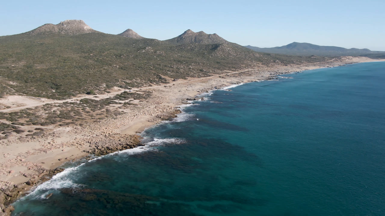 Aerial frame over Laguna Beach Los Cabos showing turquoise water, a rocky sand shoreline, scrub-covered mountains, clear sky, and no visible people.