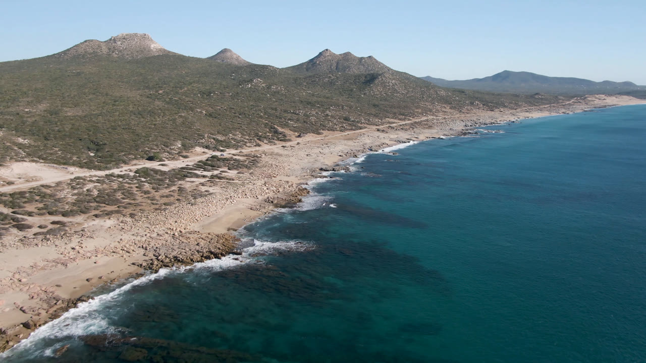Aerial view of Laguna Beach Los Cabos with darker reef-like water near shore, white wave lines, tan rocks, distant mountains, and no visible people.