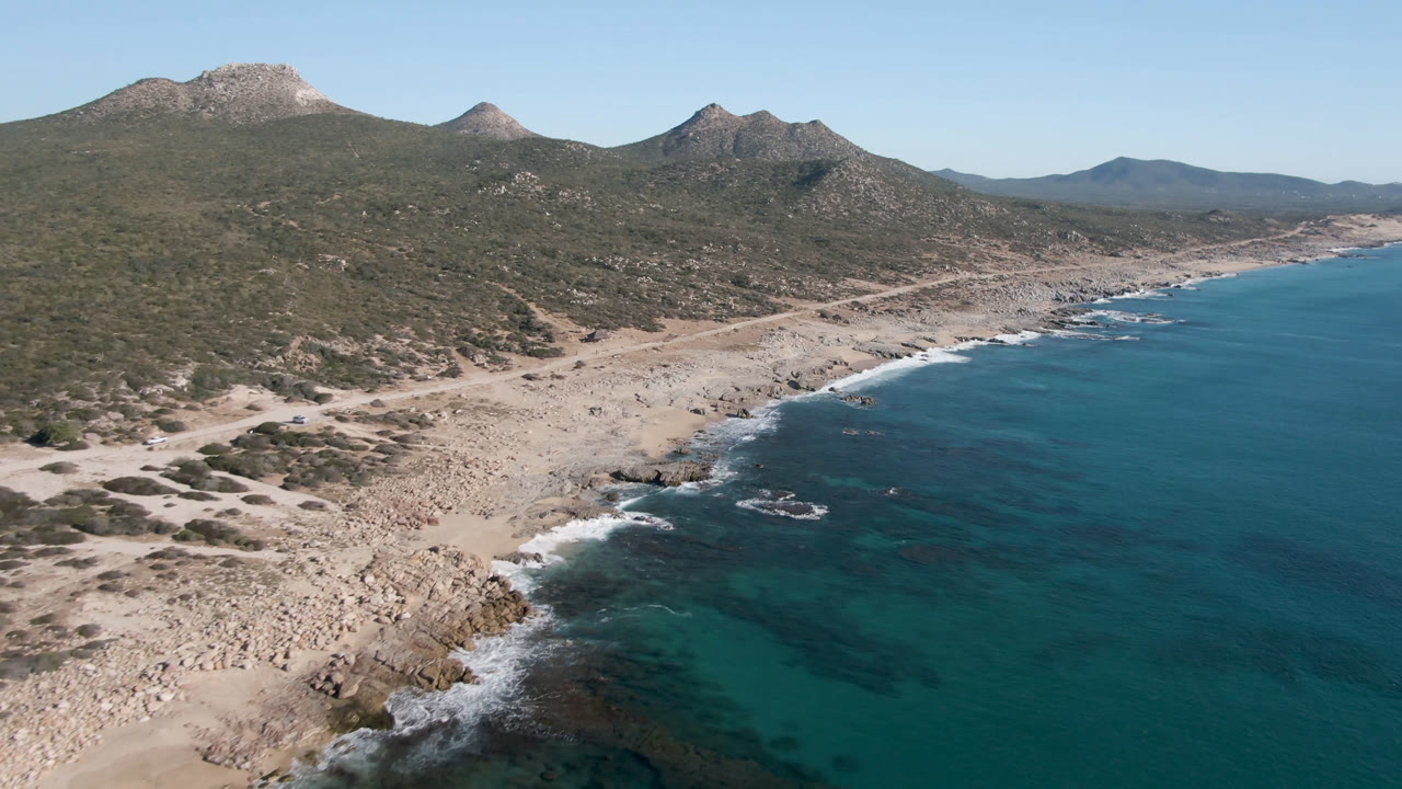 Aerial frame of Laguna Beach Los Cabos with blue ocean at right, rocky beach below, a dirt road through scrubland, mountains in the background, and no visible people.