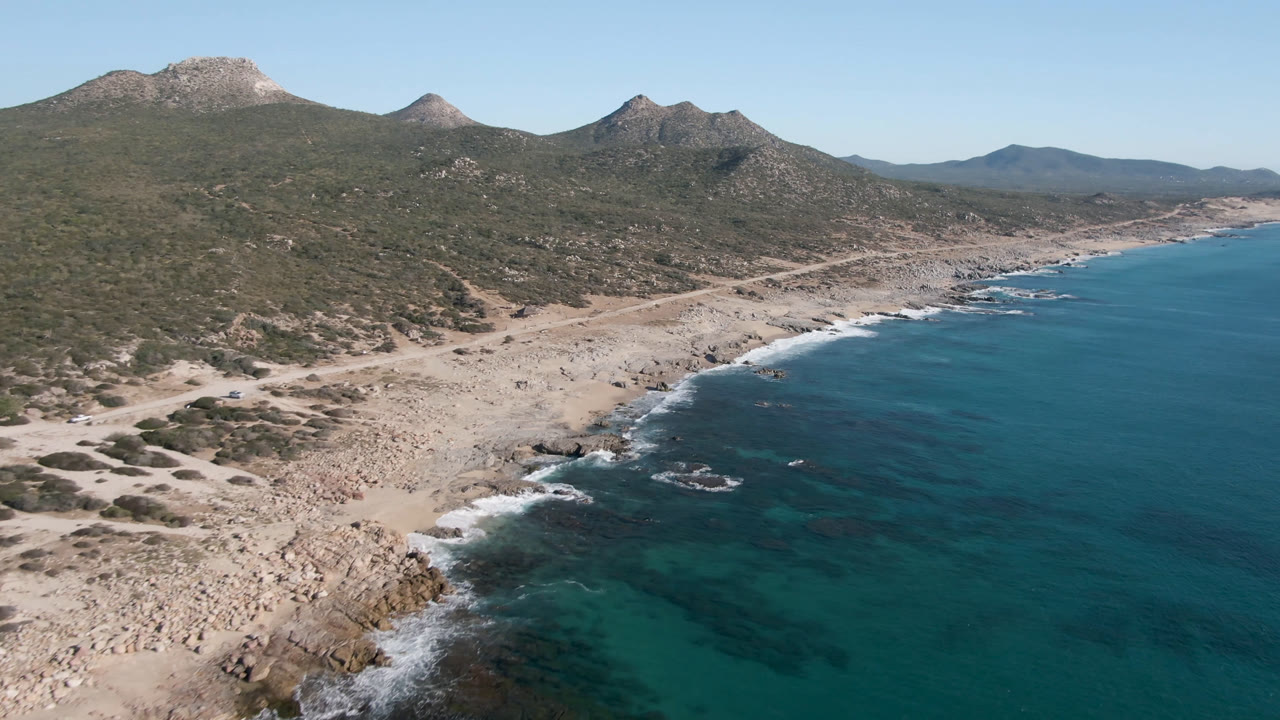 Drone view of Laguna Beach Los Cabos showing small waves on rocks, pale sand and boulders, a shoreline road, green-brown scrub hills, distant peaks, and no visible people.