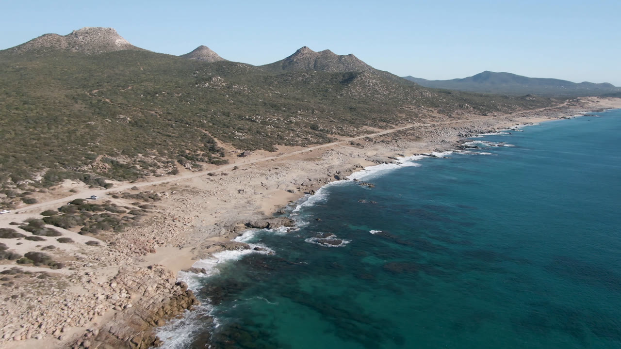 Wide coastal aerial with turquoise water, beach road, scrubland, and mountains