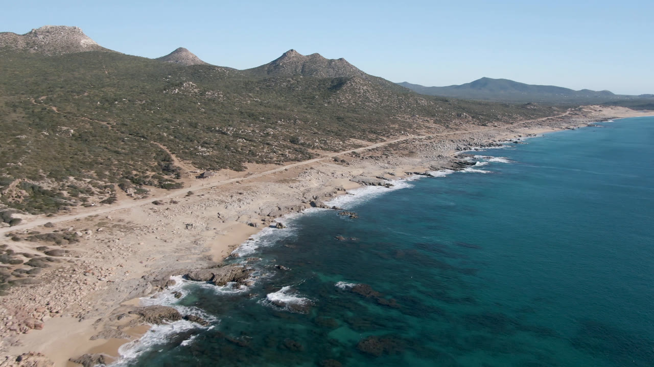 Opening aerial frame over Laguna Beach in Los Cabos, Baja California Sur, with turquoise water at right, rocky sand at left, scrub hills and mountains behind, and no visible people.