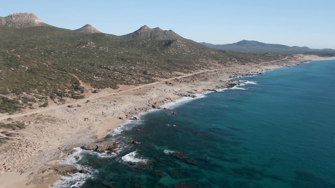 Aerial frame from Laguna Beach in Los Cabos showing small white waves around shoreline rocks, a pale dirt track near the beach, dry green-brown hills, distant mountains, and no visible people.
