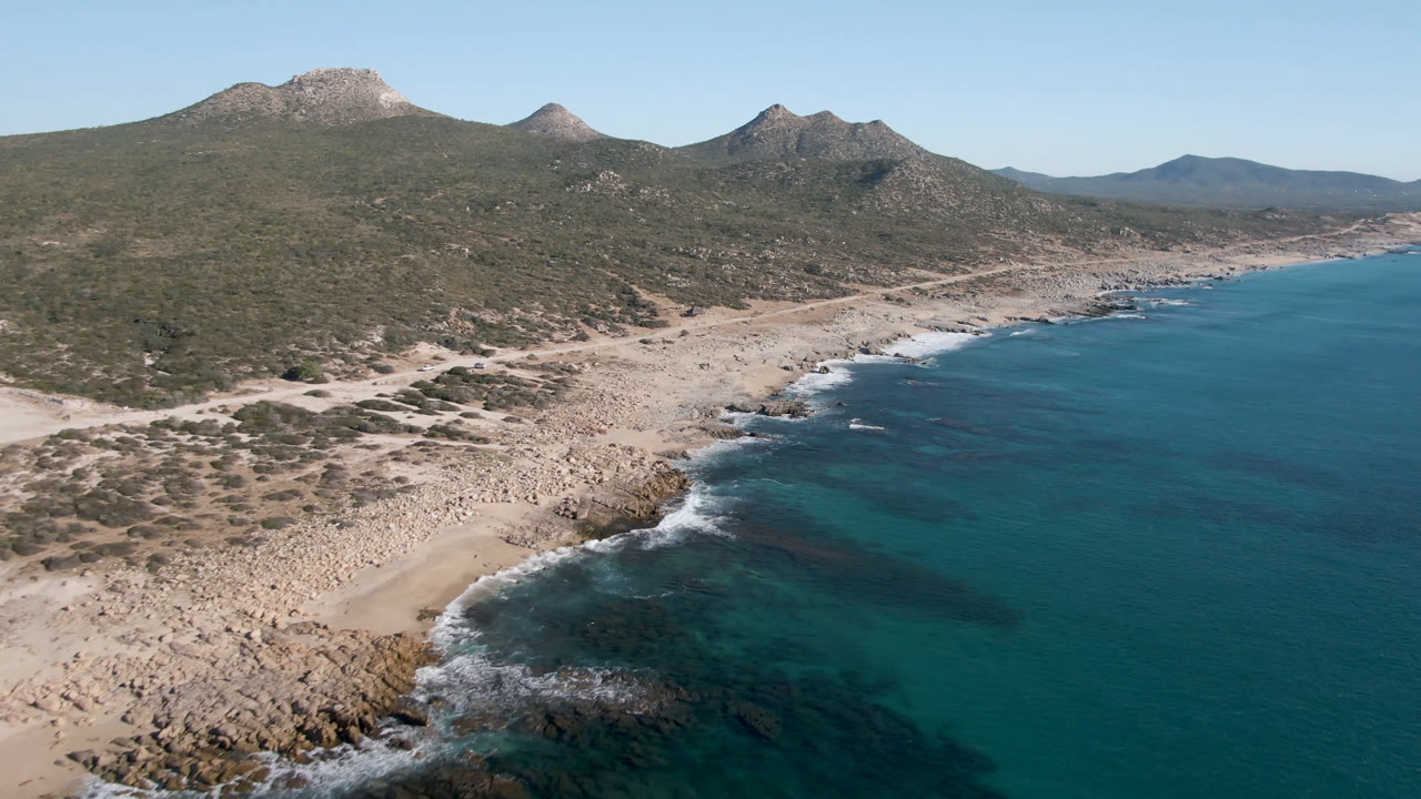 Aerial frame over Laguna Beach Los Cabos, Mexico, with turquoise water meeting a rocky sandy shore, scrub-covered hills behind it, and no visible people.