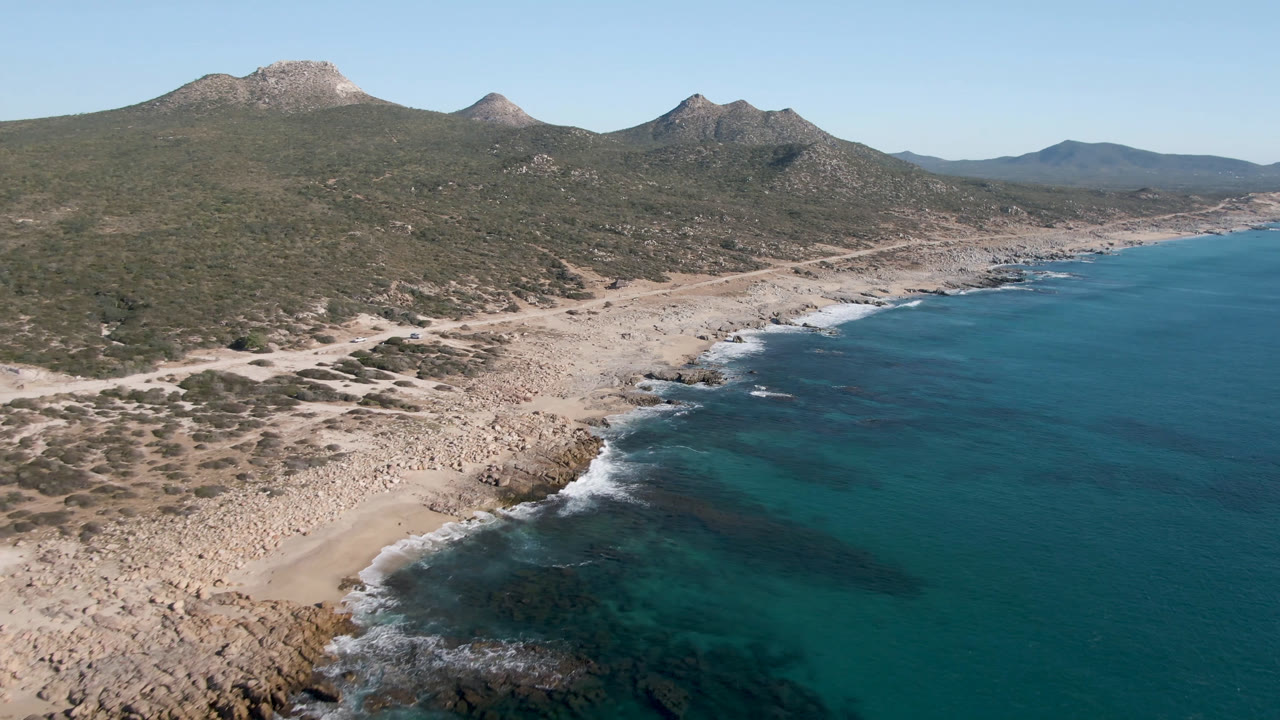 Drone view of Laguna Beach Los Cabos showing a small sandy cove below, foamy wave edges on dark rocks, open blue water, and no visible people.