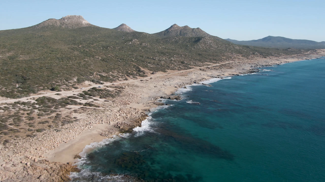 Wide aerial coastline with mountains, beach, rocks, and clear ocean.