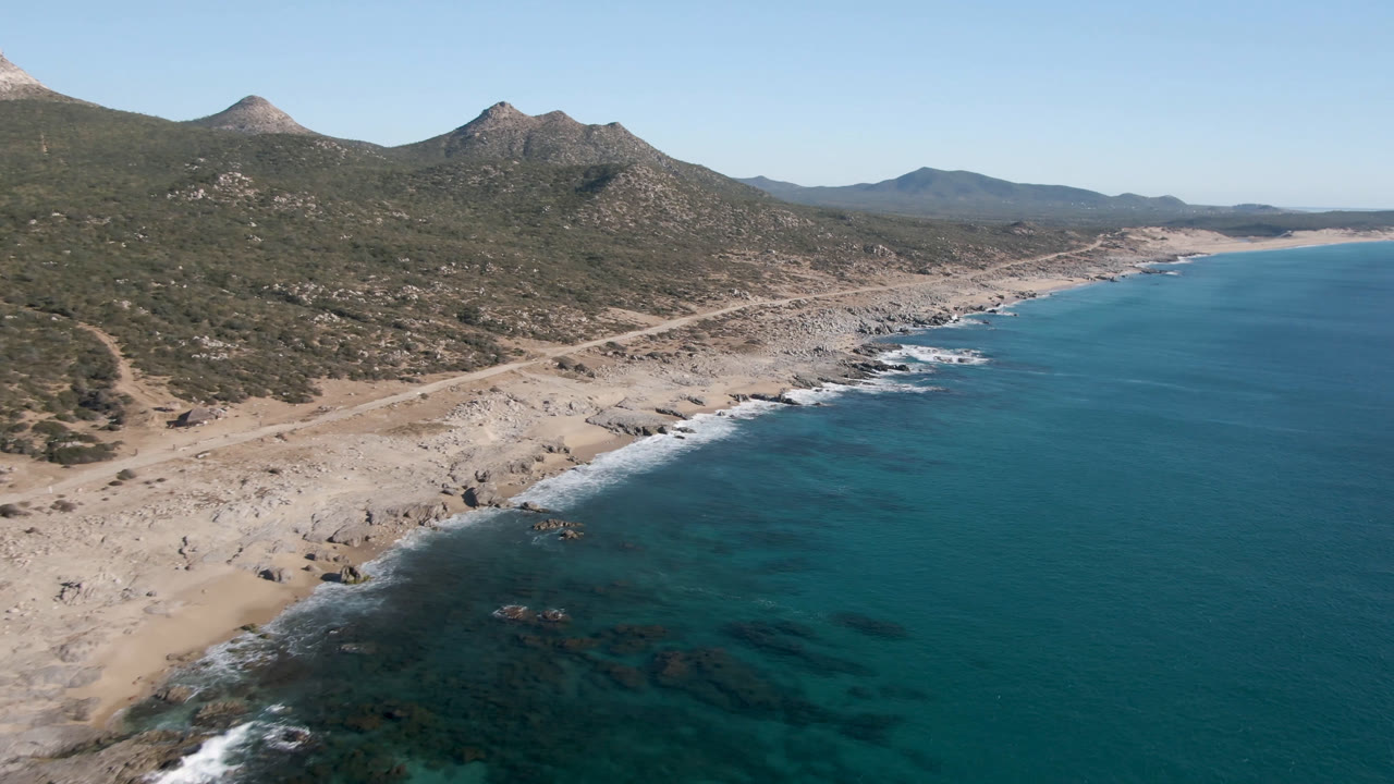Opening aerial frame over Laguna Beach in Los Cabos, Mexico, with turquoise water at right, pale rocky sand at left, scrub-covered mountains behind, and no visible people.