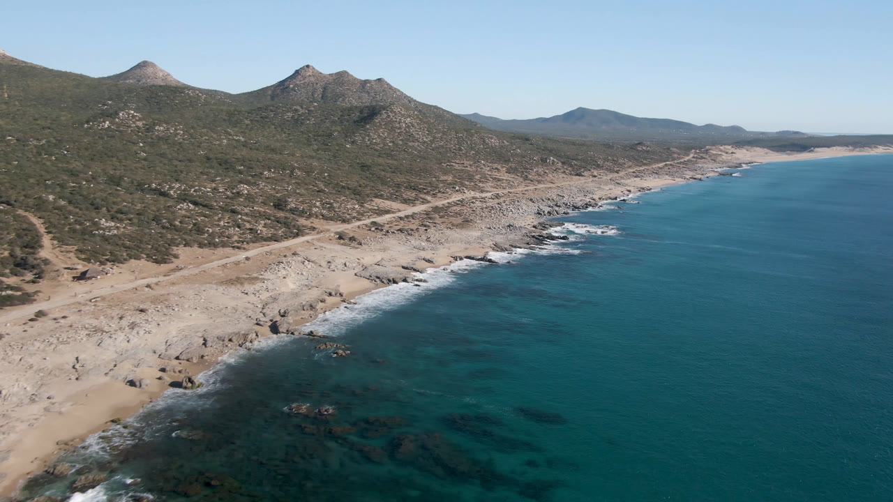 Aerial view of Laguna Beach in Los Cabos, Mexico, showing the diagonal rocky shoreline, white surf patches, open blue water, inland mountain slopes, and no visible people.