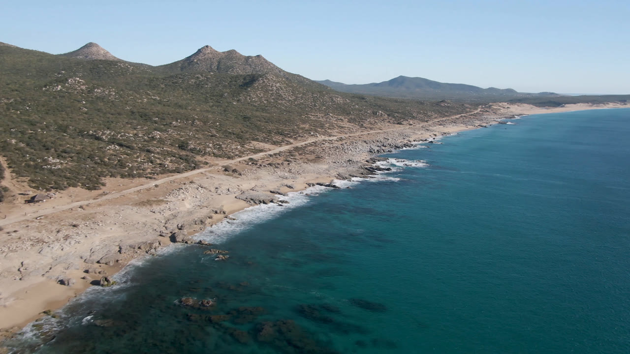 Wide aerial coastline with clear water, sandy rocks, and mountain ridges.