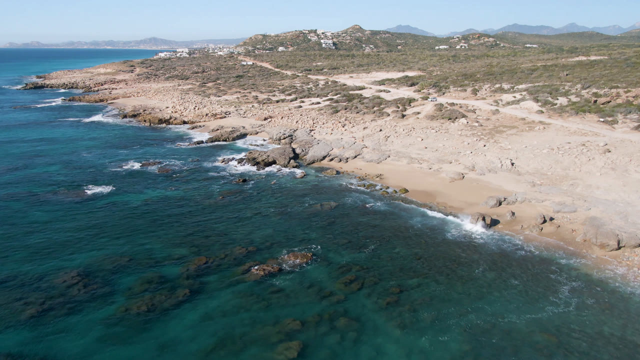Aerial frame over Laguna Beach Los Cabos showing turquoise water, a rocky sandy shore, low scrub hills, distant mountains, hillside buildings, and no visible people.