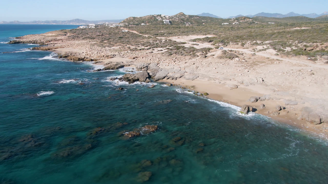 Aerial view of Laguna Beach Los Cabos with dark blue water meeting pale sand, exposed rocks near the surf line, hillside buildings in the distance, and no visible people.