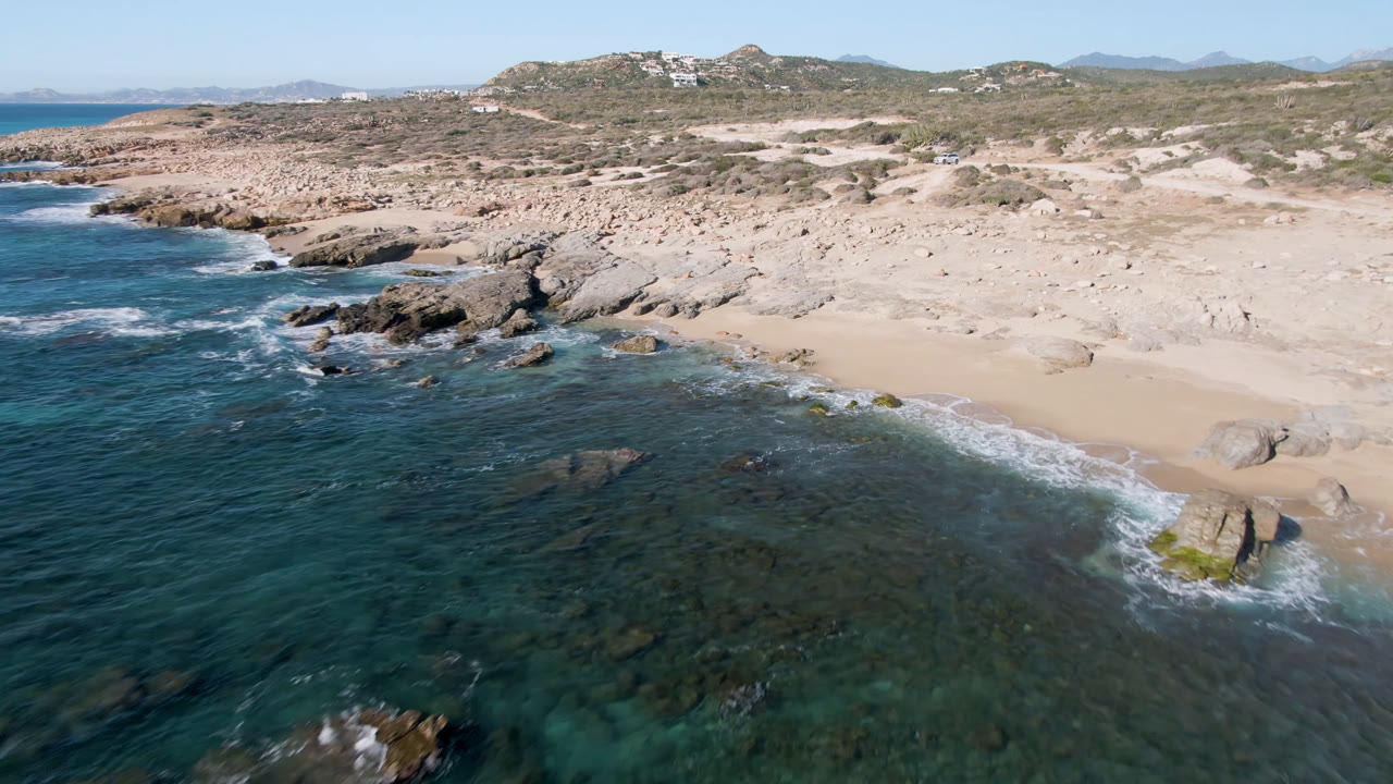 Aerial frame over Laguna Beach in Los Cabos showing clear water in the foreground, surf touching rocks and sand, dry coastal hills in back, and no visible people.