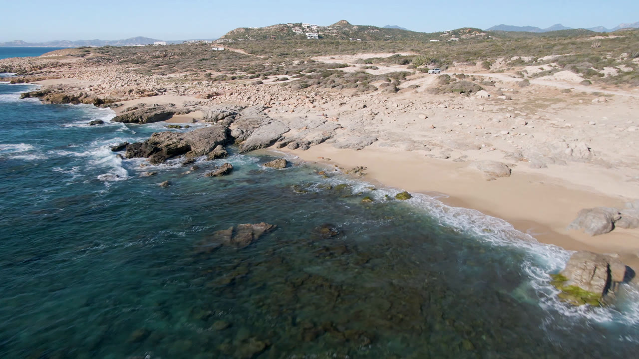 Aerial view of Laguna Beach in Los Cabos with waves spreading around dark shoreline rocks, pale sand to the right, scrubby terrain beyond, and no visible people.
