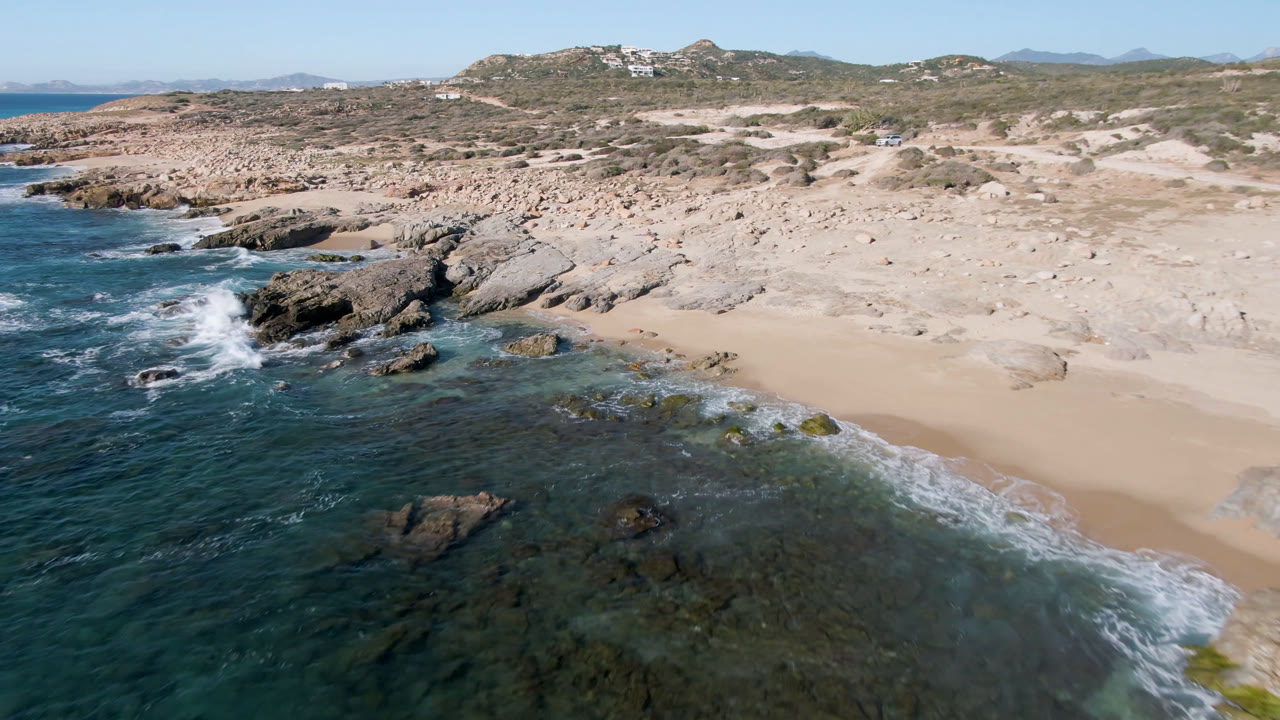 Aerial coastline with surf breaking over rocks and empty sand.
