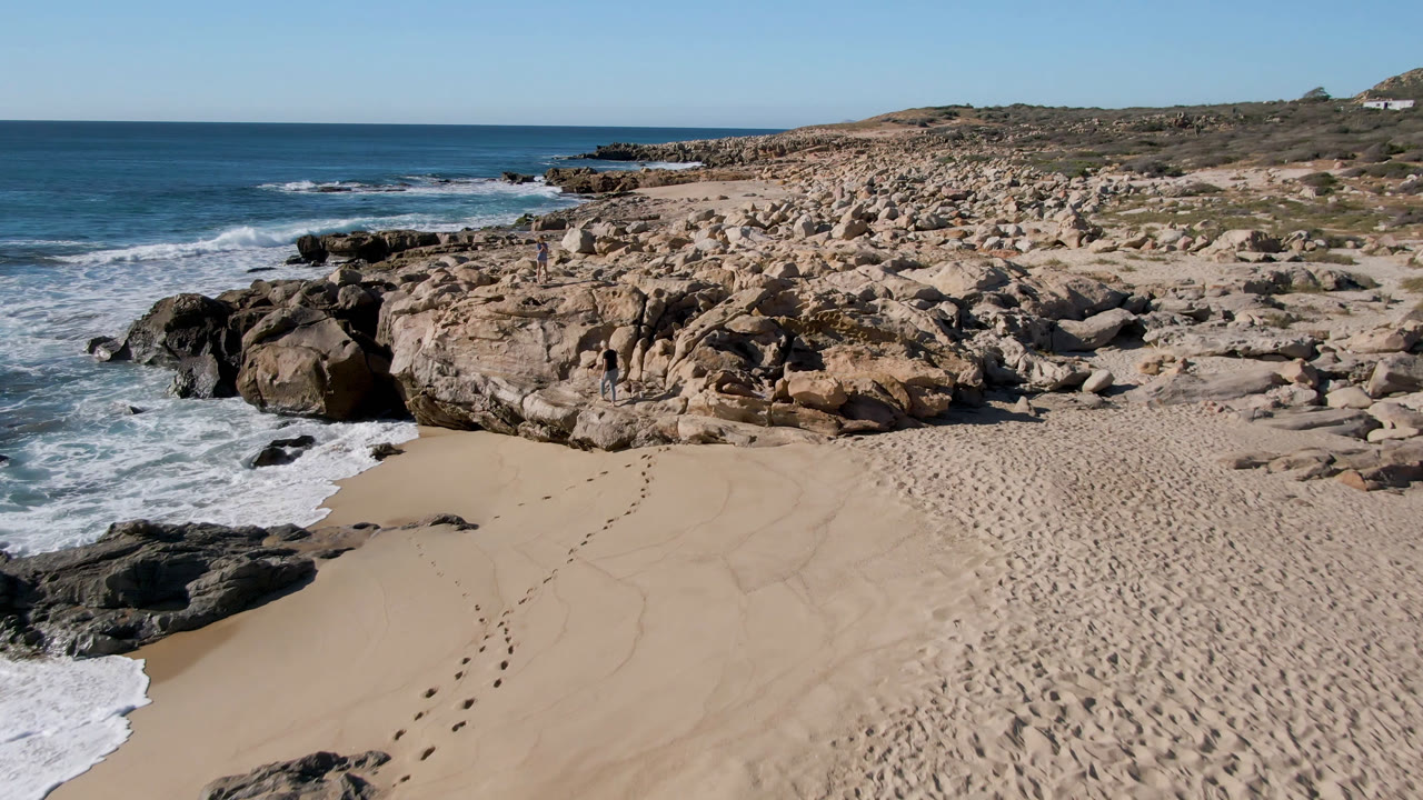 Opening frame of Laguna Beach in San Jose Del Cabo, Mexico, showing footprints across pale sand, surf entering from the left, rocky shoreline ahead, blue ocean beyond, and no visible people.