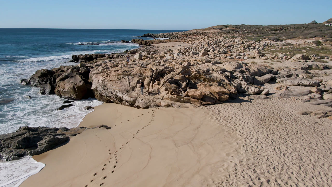 One second into the clip, footprints lead through the lower sand toward rocks while white water wraps the left edge of Laguna Beach in San Jose Del Cabo, Mexico, with no visible people.