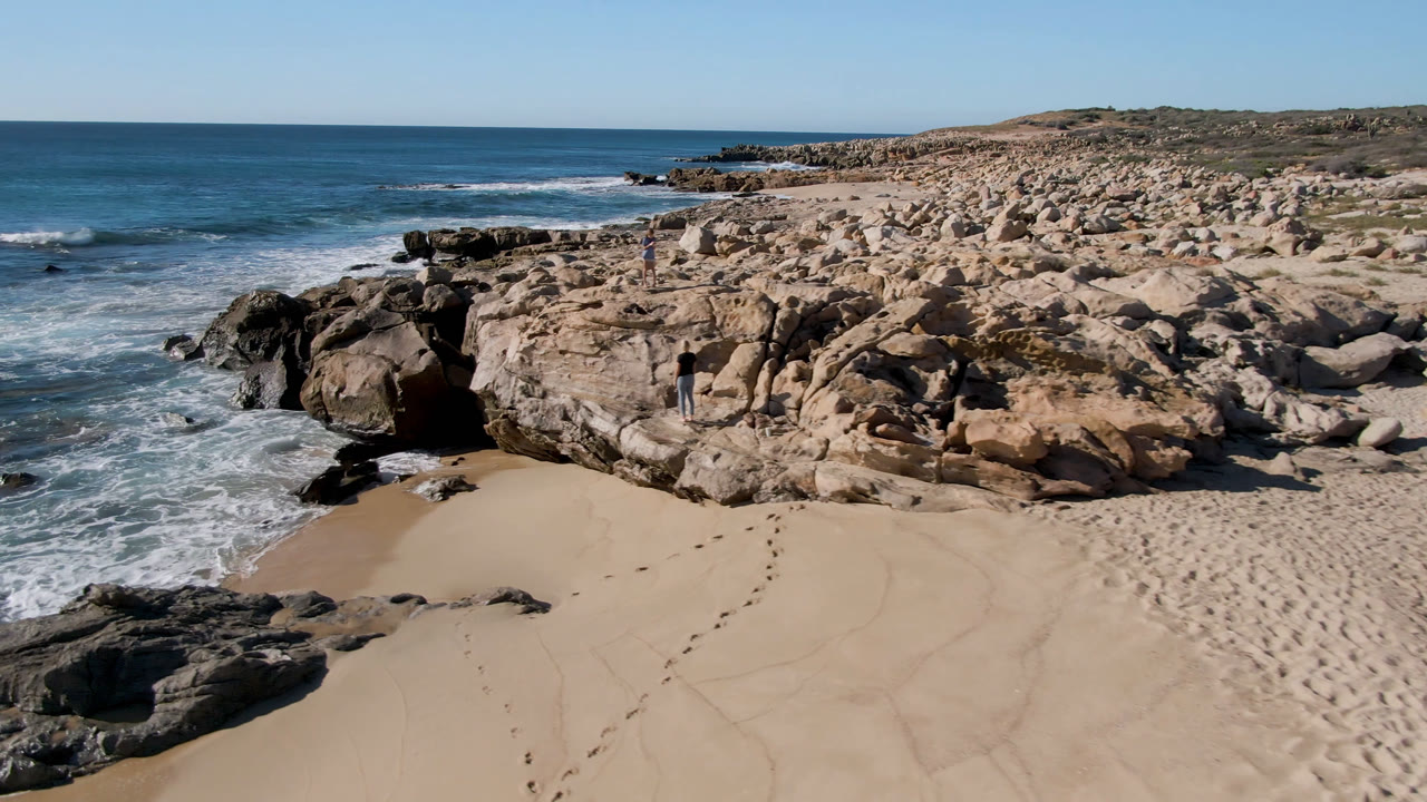 Aerial view of a sandy cove, rocky shoreline, blue ocean, and clear sky.
