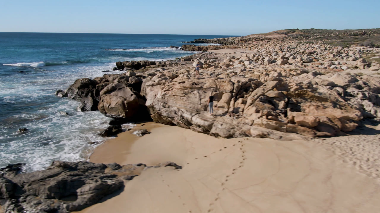 Aerial frame from San Jose Del Cabo, Mexico, with foamy surf curling along dark rocks, tan sand in the lower frame, pale boulders across the right side, and tiny distant figures on the formation.